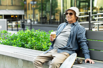 Obraz premium Elderly man with a longboard sitting on the bench and drinking coffee in a city park