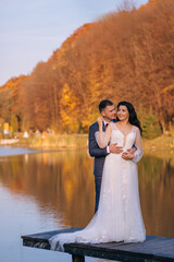 A gentle bride in a white wedding dress and a long veil, and the groom in a suit hugs a woman from behind against the backdrop of a lake, a river. Reflections of orange trees with leaves.