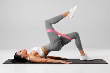 Athletic girl doing glute bridge exercise with resistance band on gray background. Fitness woman working out