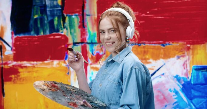 Portrait of young beautiful woman in white headphones standing near painting with artistic instruments and looking at camera smiling.