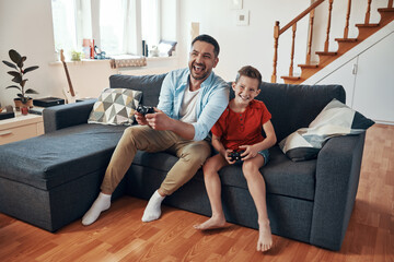 Happy young father and son playing video games while spending time at home - obrazy, fototapety, plakaty