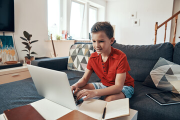 Concentrated boy doing homework using laptop while homeschooling in the living room