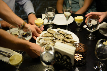 Hands putting plate with many oysters on table. People sitting by table,  with glasses and candle. - Powered by Adobe