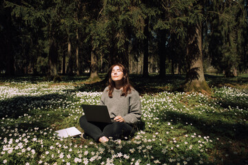 Woman with a laptop in the park. Young female sitting in grass with flowers work remotely in...