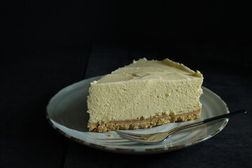 Close-up of a tasty piece of home-made lemon cheesecake and a fork on a plate with a dark background. Home baking concept with selective focus and copy space.