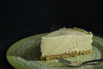 Close-up of a tasty piece of home-made lemon cheesecake and a fork on a green and white plate with a dark background. Home baking concept with selective focus and copy space.