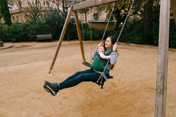 Smiling mom with little laughing baby in kangaroo backpack swinging on a swing in the park