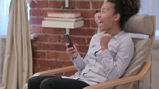 African Woman Celebrating Success on Smartphone at Home