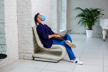 A man in a medical mask, jacket and sneakers, working on a laptop.