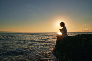 Silhouette of Peaceful woman meditating on a rock in the middle of a low wave sea at sunset time....