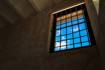 Large window with iron grating seen from inside an ancient building, blue sky and white clouds.