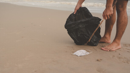 The mask has become rubbish. Right at the beach Someone is collecting and throwing away the black bag using a wooden ashtray. Behind the waves from the sea concept of garbage caused by mask