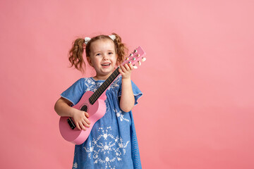cheerful little Caucasian girl of 5 years old in denim dress plays pink ukulele. child plays a musical instrument and sings on a pink background. concept of summer and the beginning of the holidays