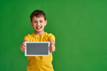 happy boy holds new computer tablet in his hands, it out in front him on green background. smiling schoolboy holds gadget with fun expression while advertising mobile device or children's learning app