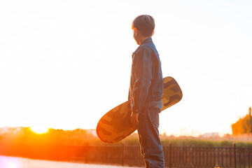 Photo of young boy holding skateboard and looking away at sunset © Vulp