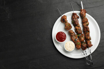 Metal skewers with delicious meat, ketchup and sauce on black table, top view. Space for text