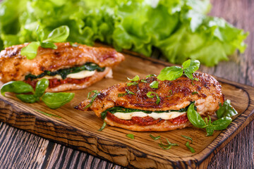 Baked chicken breast with mozzarella, sun-dried tomatoes, spinach. The meat is served on a wooden board with basil and lettuce. - obrazy, fototapety, plakaty