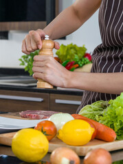 Closeup hand putting pepper on the pork before cooking it in to a steak. The kitchen counter full of various kinds of vegetables.
