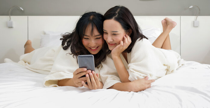 Two Asian women in bathrobe take a selfie with a mobile phone while lying in bed.