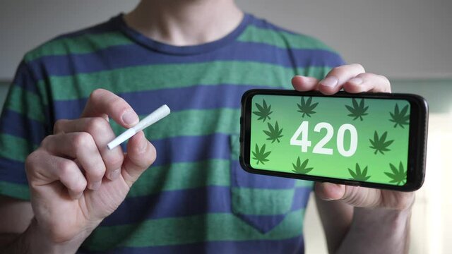 Holding a cannabis joint in one hand and a smartphone with 420 numbers on it.