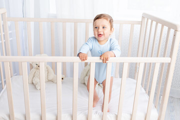 happy baby boy stands in the crib in the nursery and smiles or laughs