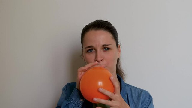 Girl prepares for holiday and inflates balloon and then lets it fly away. Young pretty woman of Caucasian European appearance inflates orange balloon with her mouth on white background.