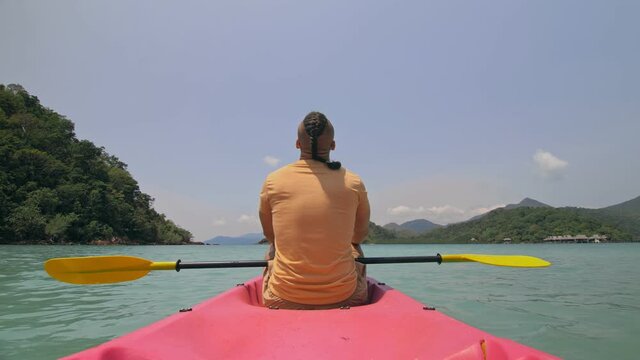 Young man with sunglasses and hat rows pink plastic canoe along sea against green hilly island with wild jungles. Traveling to tropical countries. Strong guy is sailing on kayak in ocean, back view.