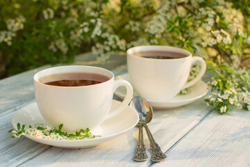 Two white porcelain cups with tea on a wooden table against a background of white flowers.