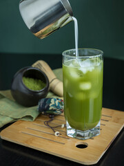 Pouring milk into a glass of iced green matcha tea .A glass of green matcha on a bamboo  tablet . A glass of  matcha tea ,a jar and a whisk on a green napking . Superfoods . Vertical orientation .