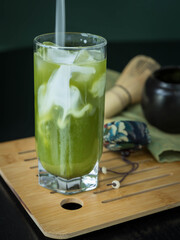 Pouring milk into a glass of iced green matcha tea .A glass of green matcha on a bamboo  tablet . A glass of  matcha tea ,a jar and a whisk on a green napking . Superfoods . Vertical orientation .
