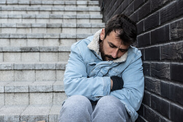 Young despair unhappy sick depressed homeless man sleeps on the steps near the sidewalk in a city street frozen and hungry suffering from depression and anxiety, social documentary concept