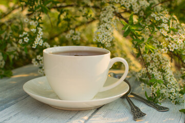 White porcelain cup with tea on a wooden table against a background of white flowers.
