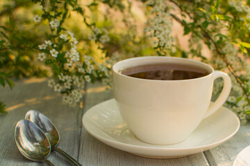 White porcelain cup with tea on a wooden table against a background of white flowers.
