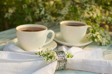 White porcelain cup with tea on a wooden table against a background of white flowers. In the foreground is a white linen napkin with a silver serving ring.