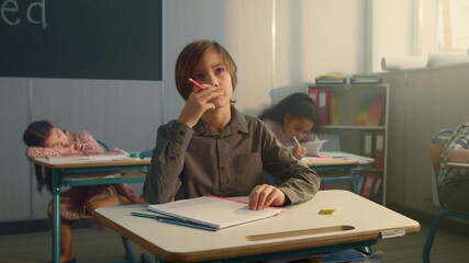 Boy sitting at desks in school class. Pensive schoolboy holding pencil in hand