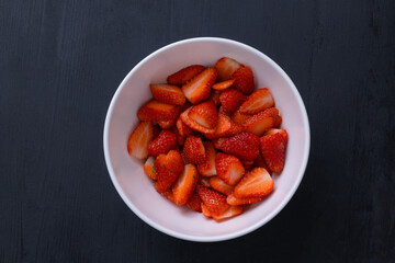 Top view picture of red fresh strawberry in a white bowl. The strawberry is cut in small piece and are covered in sugar. The bowl is on a black wooden table.