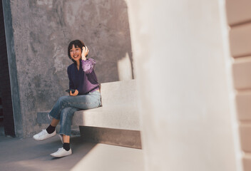 Happy young asian woman listening to music with headphones via smartphone and having fun while sitting side the street.