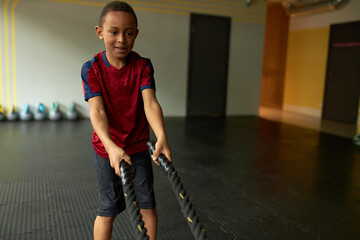 Active energetic Afro American boy wearing comfy sports clothing holding two ropes during intensive physical training, working out core and arms muscles . Activity, motivation and workout concept