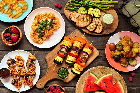 Healthy plant based summer bbq table scene. Top view on a dark wood background. Grilled fruit and vegetables, skewers, cauliflower steak and vegetarian sides. Meat substitute concept.