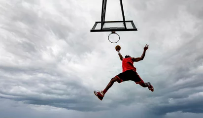 Keuken spatwand met foto Street basketball player making a powerful slam dunk on the court - Athletic male training outdoor on a cloudy sky background - Sport and competition concept © Davide Angelini