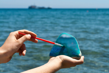 hands with stone and paintbrush in front of blue sea