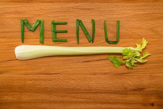 Onion petals folded in the form of the word 'MENU' and celery on wooden table. Close-up view from above