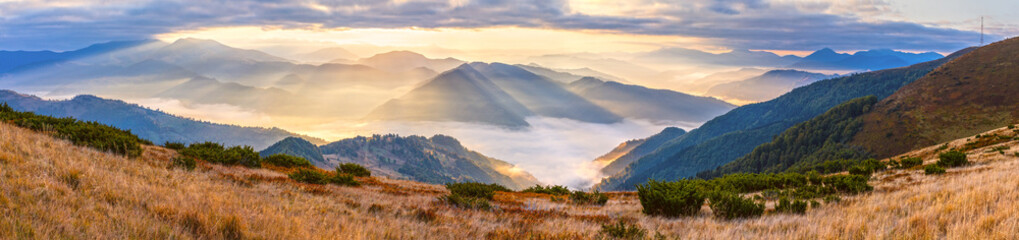 Autumn morning mountain panoramic view with sunbeams through haze and low clouds.
