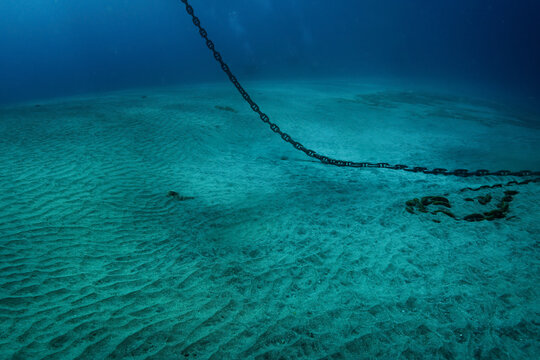 Underwater photo of heavy steel anchor chain that hangs down from water surface and lays on white sand bottom, murky blue water deeper in tropical sea