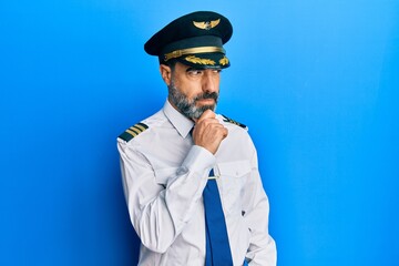 Middle age man with beard and grey hair wearing airplane pilot uniform with hand on chin thinking about question, pensive expression. smiling with thoughtful face. doubt concept.