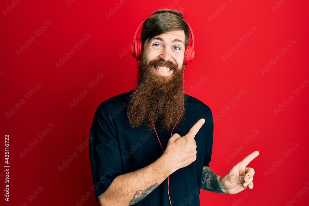 Poster Redhead man with long beard listening to music using headphones smiling and looking at the camera pointing with two hands and fingers to the side.