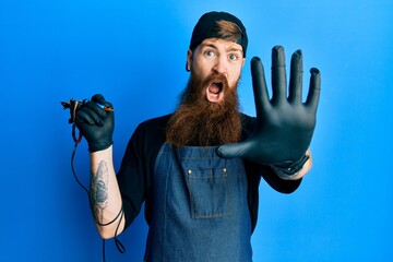 Redhead man with long beard tattoo artist wearing professional uniform and gloves doing stop...
