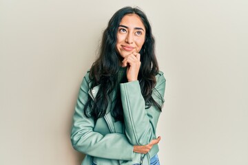 Hispanic teenager girl with dental braces wearing green leather jacket thinking concentrated about...