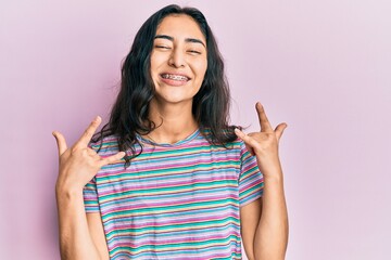 Hispanic teenager girl with dental braces wearing casual clothes shouting with crazy expression doing rock symbol with hands up. music star. heavy concept.