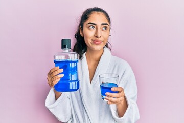 Hispanic teenager girl with dental braces holding mouthwash smiling looking to the side and staring away thinking.